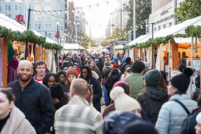 Shoppers walking between decorated wooden vendor stalls under strings of lights at an outdoor holiday market
